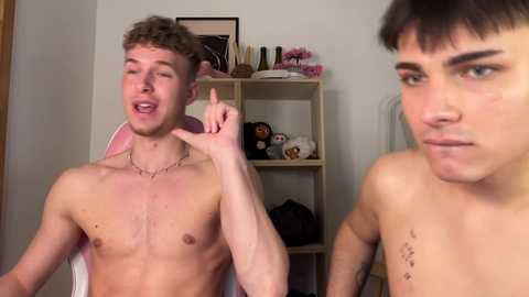 Video of two shirtless young men in a bedroom; one with curly hair and a necklace, the other with straight hair, both making playful faces.