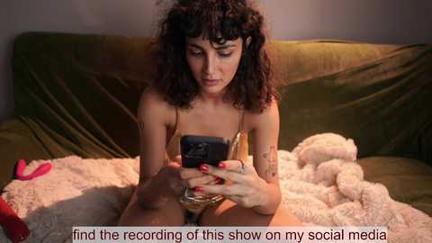 A video shows a dark-haired woman with tattoos, wearing a bra, sitting on a bed, engrossed in her smartphone. A red vibrator is beside her.