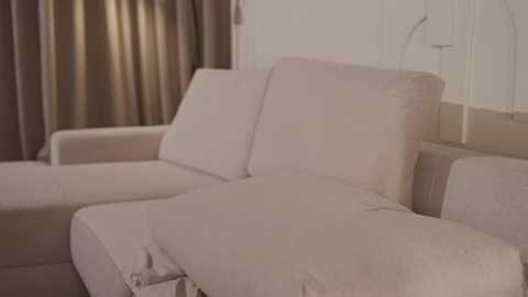 Video of a beige sectional sofa with light beige cushions in a minimalist living room with beige curtains and white walls. The setting is warm and cozy.