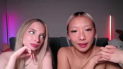 Video of two topless young women with fair skin and blonde hair, one with a thoughtful expression, the other smiling. Background shows a gray couch, red and purple lights, and a stuffed animal.