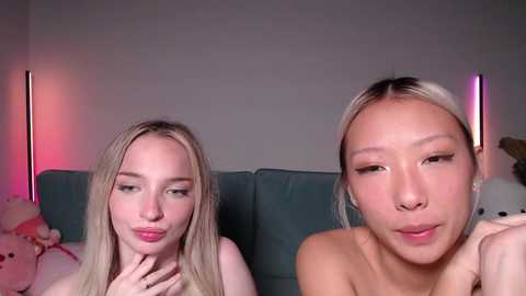 Video of two young women, one blonde and one with black hair, on a teal couch, both topless, smiling, in a dimly lit room with pink and purple lights.