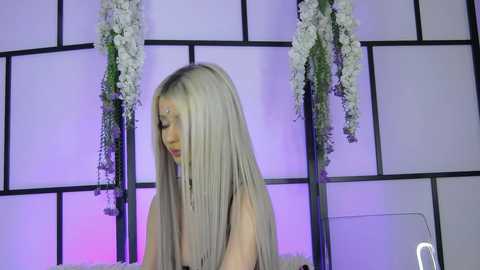 Media: Video of a fair-skinned woman with long, straight, platinum blonde hair, wearing a sleeveless black dress, in front of a shoji screen adorned with white flowers.