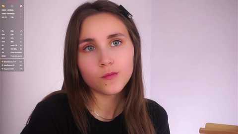 Video of a young woman with fair skin, blue eyes, straight brown hair, and a black hair clip. She wears a black top, and the background features a white wall and a black clock.