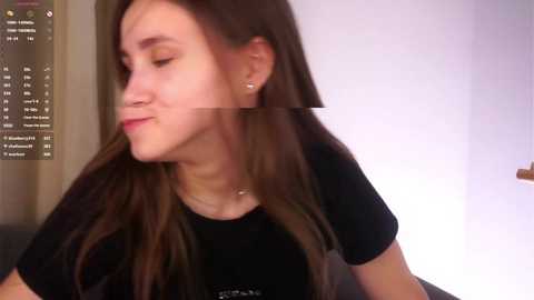 A video of a young woman with long brown hair, wearing a black T-shirt, and a necklace, smiling slightly, with a computer screen in the background.