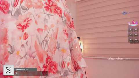 Video of a woman wearing a pink and white floral dress, standing beside a wall with a mounted TV. The room has a modern, minimalist decor with white and beige tones.