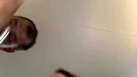 A video of a man with short brown hair, wearing a red shirt, holding a metal object, taken from a low angle. The background is a plain, off-white wall.