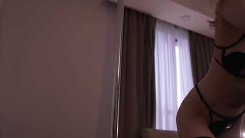 A dimly lit video captures a woman in black lingerie, standing beside a mirrored wardrobe. She has a slender physique, with her breasts partially visible. The background features a window with sheer curtains and dark drapes. The room is softly lit.
