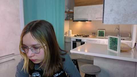 Video of a young girl with long blonde hair, glasses, and a black shirt, seated in a modern kitchen with white cabinets, teal accents, and a green wall art.