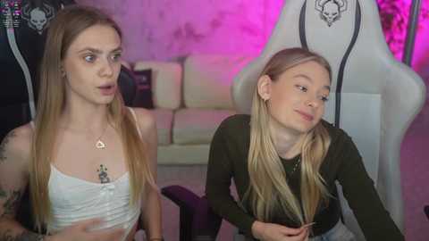 Media: Video of two young women, one with long blonde hair in a white crop top, the other with long blonde hair in a green long-sleeve top, sitting in a gaming room with a white gaming chair and purple lighting.