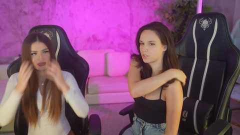 Media: Video of two young women, one with long brown hair and a white top, and the other with medium brown hair and a black tank top, seated in black gaming chairs in a dimly lit room with purple lighting and a white couch in the background.