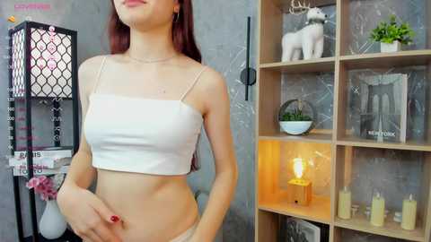 Video of a slender, fair-skinned woman with long red hair, wearing a white crop top and white panties, standing in a modern, minimalist room with wooden shelves and a lit candle.