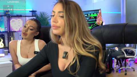 Video of two women in a modern office setting. One, with long blonde hair, wears a black top, appears in the foreground. The other, with dark hair, in a white tank top, is seated behind.