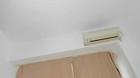 Media: Video of a ceiling with a white air conditioning unit and beige curtains covering a window. The room has plain white walls, and the ceiling appears flat and smooth.