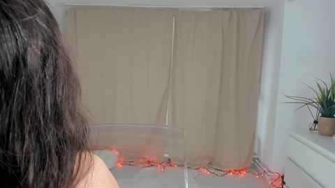 Video of a woman with long, dark hair, standing with her back to the camera, looking at a beige curtain and a white window sill adorned with small potted plants and fairy lights.