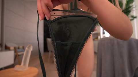 A close-up video of a woman holding a black lace thong, her hand visible in the frame, taken in a modern, brightly-lit room with a wooden floor.