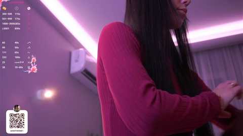 Video of an Asian woman with long black hair, wearing a red sweater, in a modern room with purple lighting. A digital clock and QR code are visible in the upper left corner.