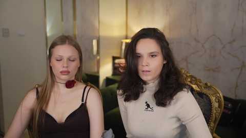Video of two women, one blonde in black lingerie, another brunette in beige turtleneck, seated indoors with beige walls and green furniture.