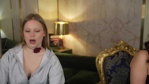 Video of a blonde woman in a gray robe with a red rose, sitting on a green velvet couch in a dimly lit room with gold accents and a lamp.