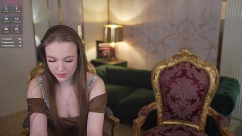 Video of a young Caucasian woman with long brown hair, wearing a brown off-shoulder top, sitting on an ornate gold and red velvet chair in a warmly lit, elegantly decorated room with a green sofa and a lamp.