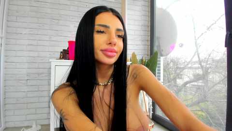 Video of a young, light-skinned woman with long black hair, wearing a gold necklace and no top, in a modern room with a brick wall, greenery, and a window.