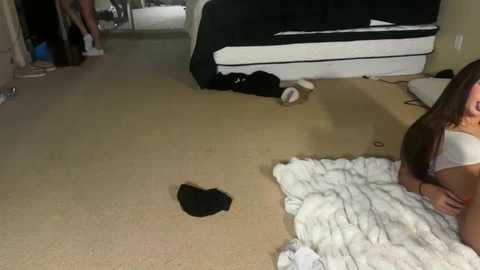 Video of a messy living room with beige carpet, a white blanket, and scattered black clothes.