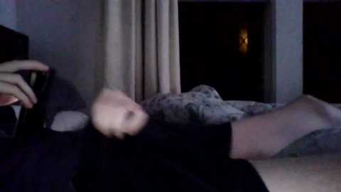 Video of a dimly lit bedroom with a person lying on a bed, partially covered by a patterned blanket. The room features beige curtains and a warm light source from a nearby lamp.