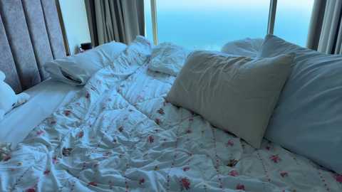 A video of a neatly made bed with white floral-patterned sheets, large white pillows, and a plush gray upholstered headboard. The background features a large window with sheer curtains, revealing a serene, blue ocean view.