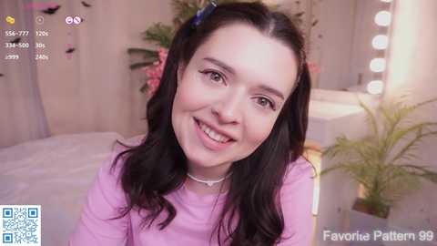 Video of a smiling young woman with long black hair, wearing a pink sweater, in a cozy, lit bedroom with a floral background.
