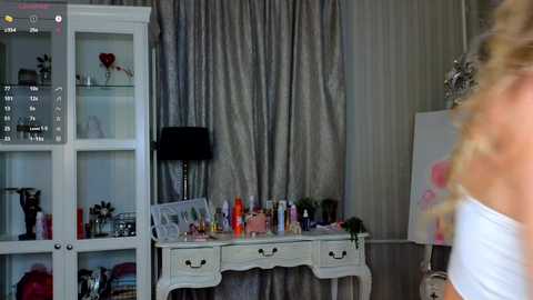 Video of a woman with blonde, wavy hair, partially visible, in a white dress, standing beside a white vanity table with a black lamp, surrounded by cosmetics and framed photos, in a room with grey curtains and a glass cabinet.