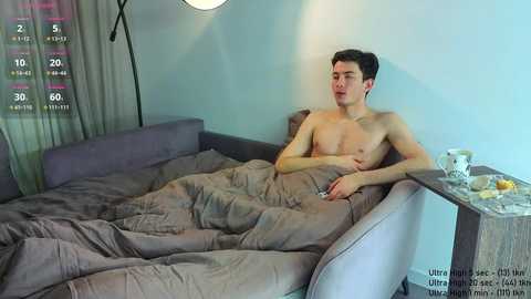 Video of a young, shirtless man with short dark hair, lying in bed under a gray duvet, in a modern, minimalist bedroom with a large lamp and a small wooden side table.