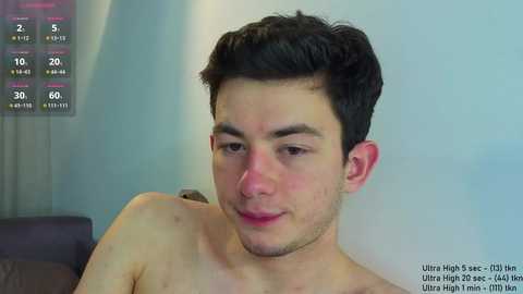Video of a shirtless young man with short dark hair, fair skin, and a slight smile, in a dimly lit room with a blurred background.
