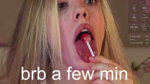 Video of a blonde woman with light skin, licking a red lollipop with white letters \"G\" and \"C.\" Background shows a clock and a blurred room. Text reads \"brib a few min.\