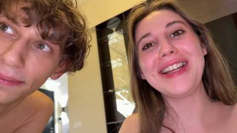 Video of a smiling young woman with light brown hair and a young man with curly hair, both looking happy in a bathroom with a mirror and white wall.