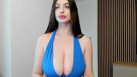 Media: Video of a fair-skinned, voluptuous woman with long dark hair, wearing a revealing blue halter top that accentuates her large breasts, standing indoors against a background of vertical wooden slats.