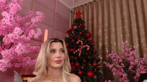 A video of a blonde woman with fair skin and a slender figure, wearing a sleeveless dress, standing next to a decorated Christmas tree adorned with red ornaments and a star topper. The background features pink cherry blossoms and beige curtains.