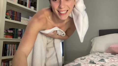 A video of a young, light-skinned woman with short brown hair, wearing a white towel, smiling. She is in a bedroom with a bookshelf filled with books and a bed with a floral duvet.