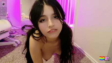 Video of a young Latina woman with long, dark hair, fair skin, and full lips, kneeling on a beige carpet in a dimly lit room with purple neon lights.