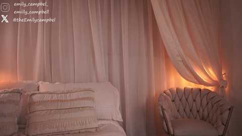 Video of a cozy, dimly lit bedroom with a cream-colored tufted chair and a white quilted bed, draped in sheer curtains with soft orange light.