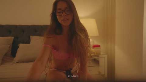 Video of a slender, fair-skinned woman with long brown hair and glasses, wearing pink lingerie, sitting on a bed in a dimly lit room.