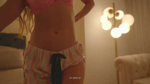A video of a woman in a pink bra and striped pajama shorts, standing in a dimly lit room with a chandelier and a beige couch.