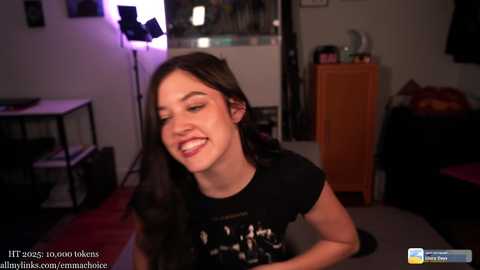 Video of a smiling young woman with long dark hair, wearing a black t-shirt, in a dimly lit room with a lamp, wooden cabinet, and couch.