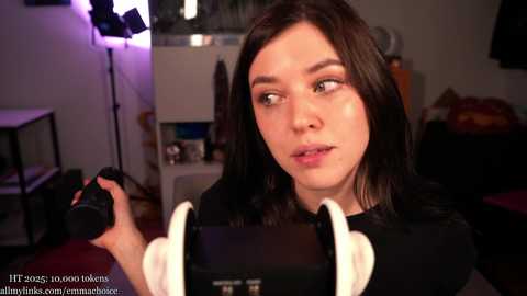 Video of a young woman with straight black hair, wearing a black shirt, holding a camera, in a dimly-lit, cluttered room with studio lights and equipment in the background.