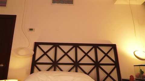 Video of a cozy bedroom with a dark wooden headboard featuring a geometric pattern. The bed is neatly made with white linens. A modern, crescent-shaped floor lamp casts a warm glow on the right side. The walls are beige with a vent near the ceiling.