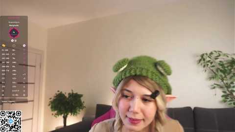 Media: Video of a young woman with light skin, blonde hair, and elf ears, wearing a green knitted hat with ears, in a cozy living room with a calendar, plants, and a grey couch.