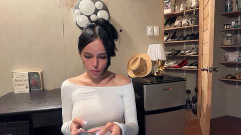 Video of a woman with dark hair in a bun, wearing a white off-shoulder top, examining a phone in a cluttered, beige-walled room with a wooden shelf filled with shoes and a hat, and a table holding a book and lamp.