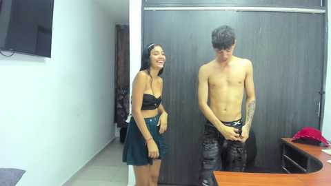 Video of a young woman with long hair in a black strapless top and green skirt, and a shirtless man with tattoos in camouflage pants, both laughing in a dimly lit room.