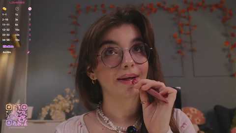 A video of a young woman with fair skin and shoulder-length dark hair, wearing large round glasses, a necklace, and a pink top, biting her finger while in a dimly lit room with a colorful, festive background.