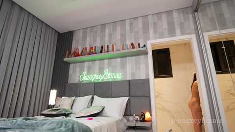Media: Video of a modern, minimalist bedroom with a gray upholstered headboard, a neon sign reading \"Champagne Sunrise,\" a shelf of colorful vases, and a partially open door revealing a bathroom with marble walls.