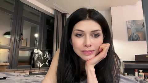 A video of a young woman with long black hair, light skin, and dark eyes, lying on a bed in a modern bedroom with a large window, abstract art, and a black lamp.