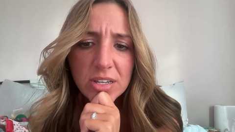 A close-up video of a distressed woman with long, wavy blonde hair, wearing a ring, with a furrowed brow and clenched teeth, sitting in a cluttered bedroom with white walls and a bed.